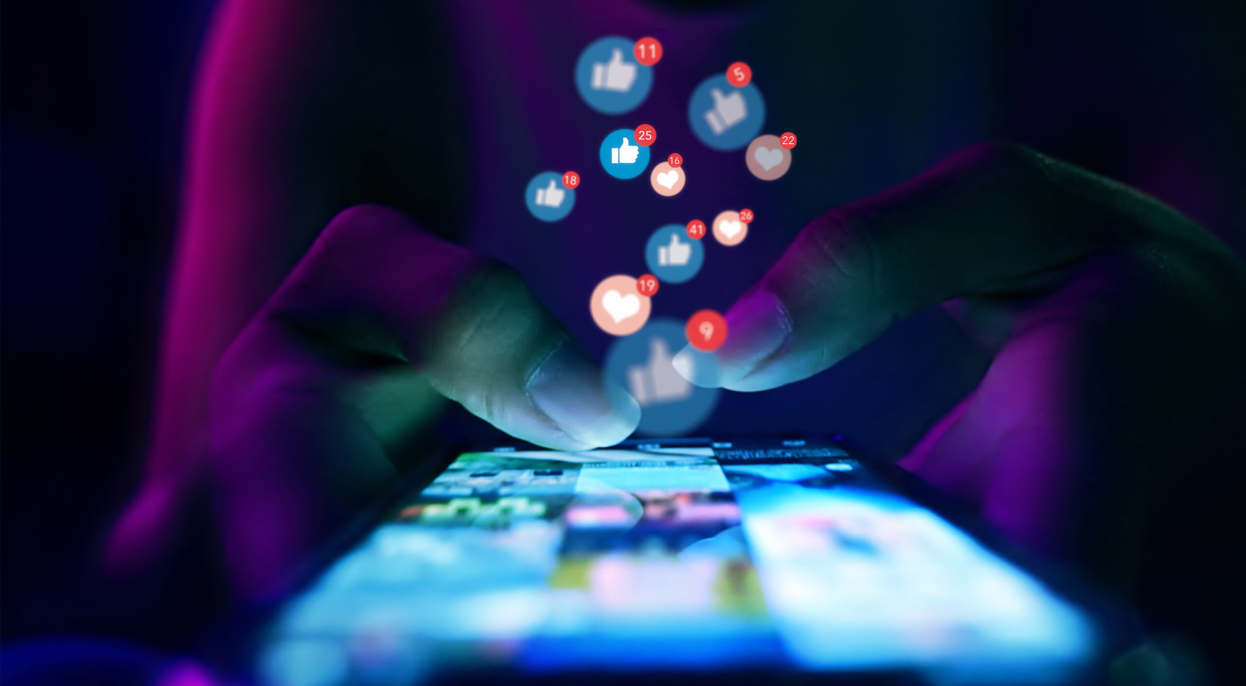 A closeup of hands touch a phone while bubbles with thumbs up and hearts float upward, suggesting the person is using social media