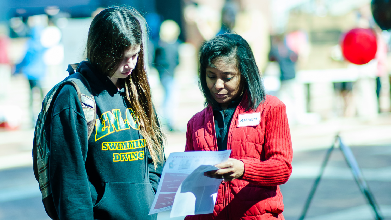 Mardecia stands on campus holding a piece of paper while talking to a student and pointing something out on the paper