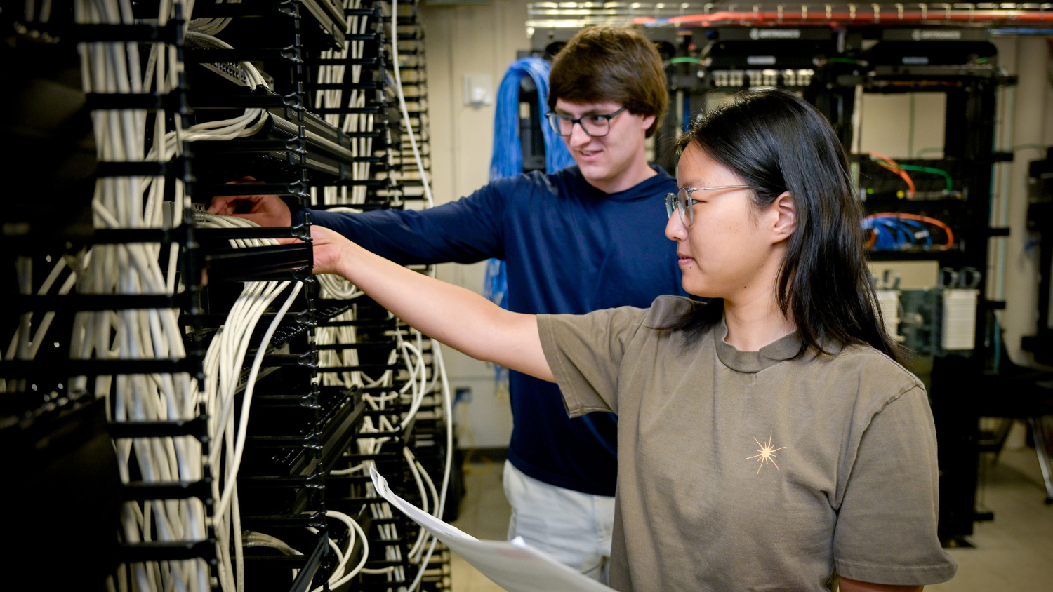 Two students reach toward cables in a networking closet.